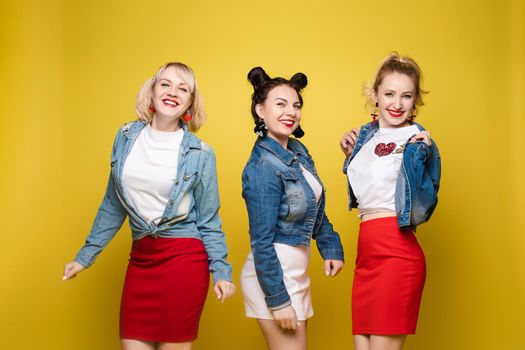 Front view of bright three girls in white shirts keeping balloons and ice creams on yellow isolated background. Cheerful pretty women looking at camera and posing. Concept of party and sparkles.