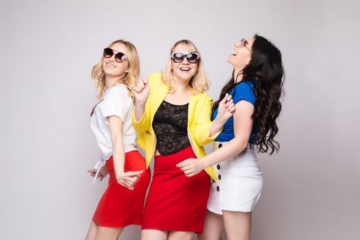 Three cheerful women wearing bright colorful skirts, shirts and glasses standing on grey isolated background. Happy girls looking at camera, laughing and showing tongue. Concept of happiness.