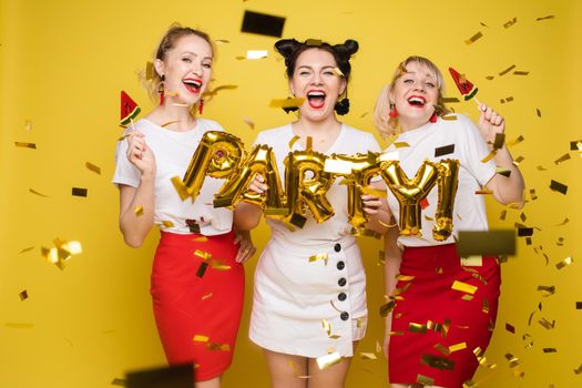 Front view of bright three girls in white shirts keeping balloons and ice creams on yellow isolated background. Cheerful pretty women looking at camera and posing. Concept of party and sparkles.