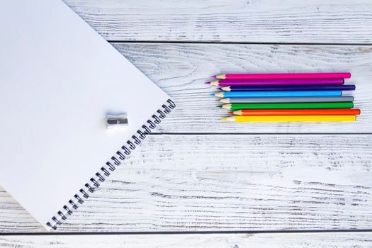 Bright, multicolored pencils for drawing in an album, a metal sharpener and a notebook with white sheets and a black spiral on a light, gray and wooden background.