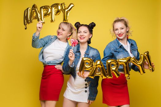 Front view of bright three girls in white shirts keeping balloons and ice creams on yellow isolated background. Cheerful pretty women looking at camera and posing. Concept of party and sparkles.