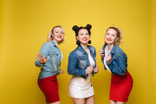 Front view of bright three girls in white shirts keeping balloons and ice creams on yellow isolated background. Cheerful pretty women looking at camera and posing. Concept of party and sparkles.