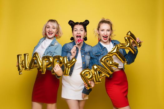 Front view of bright three girls in white shirts keeping balloons and ice creams on yellow isolated background. Cheerful pretty women looking at camera and posing. Concept of party and sparkles.