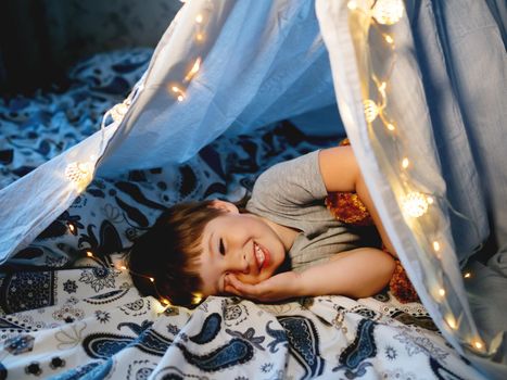 Little boy plays with his teddy bear. Toddler lying in tent made of linen sheet on bed. Cozy evening with favorite toy in DIY interior.