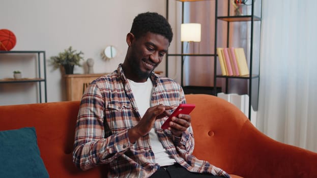 Joyful African American young man in casual shirt sitting on sofa, using smartphone or mobile cell phone in modern room at home. Tapping, scrolling, watching video, blogger content, online shopping