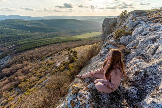 A girl sits on the edge of the cliff and looking at the sun valley.Woman enjoying a sunset or sunrise from the top of the mountain in summer.The traveler girl see the sunrise at the peak of mountain
