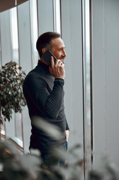 Adult man chatting on smartphone while standing near big window in the office