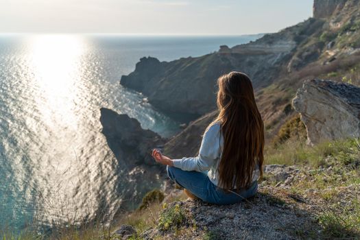 Woman tourist enjoying the sunset over the sea mountain landscape. Sits outdoors on a rock above the sea. She is wearing jeans and a blue hoodie. Healthy lifestyle, harmony and meditation.