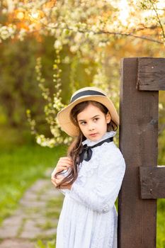 A romantic brunette little girl in a hat and white dress stands near a wooden fence, under flowering trees in spring. Vertical