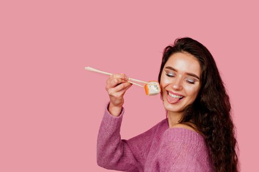 Happy girl with sushi on pink background. Young girl is holding sushi by chopsticks and wanting to eat one piece of roll philadelphia. Sushi delivery.