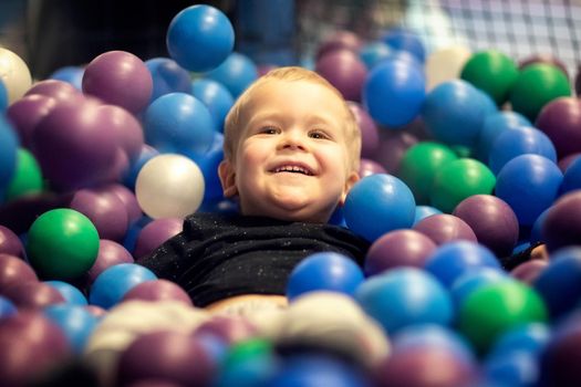 A boy lies among multi-colored plastic balls in a playroom. Happy Birthday party. Happy little kid boy playing at colorful plastic balls playground