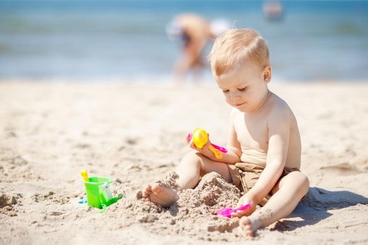 Kid playing on beach with sand. Sand and water toys. No sun protection for young child. Little boy digging sand, building castle at sea shore.