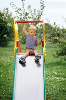 Little boy siting on a slide and contemplating to slide down. Green nature background. Childhood joys in summer time