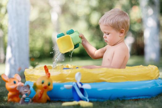 Child is sitting in a pool at the garden and playing with water toys. Hot summer day in nature. Blue yellow pool full of toys.