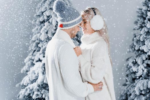 Happy young couple hugs and smiles and snow falls near christmas trees at the eve of new year celebration in winter day. Smiley man and woman weared white pullovers love each other