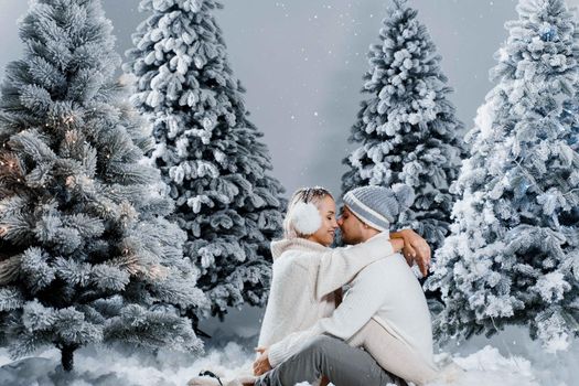 Winter love story with couple weared fur headphones, hats, white sweaters. Happy young couple hugs and kiss near christmas trees at the eve of new year celebration in winter day.