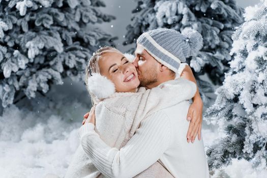 Winter love story with couple weared fur headphones, hats, white sweaters. Happy young couple hugs and kiss near christmas trees at the eve of new year celebration in winter day.