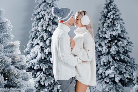 Winter love story with couple weared fur headphones, hats, white sweaters. Happy young couple hugs and kiss near christmas trees at the eve of new year celebration in winter day.