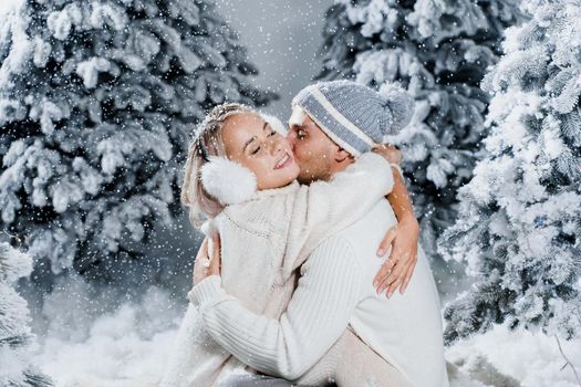 Falling snow and kisses. Happy young couple close-up hugs and kiss near christmas trees at the eve of new year celebration in winter day. Smiley man and woman weared white pullovers love each other