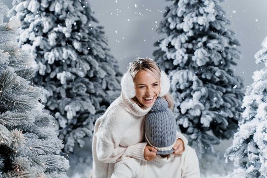 Falling snow and kisses. Happy young couple close-up hugs and kiss near christmas trees at the eve of new year celebration in winter day. Smiley man and woman weared white pullovers love each other