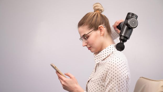 A young woman sits on a chair, uses a smartphone and makes herself massage with an electro massage gun