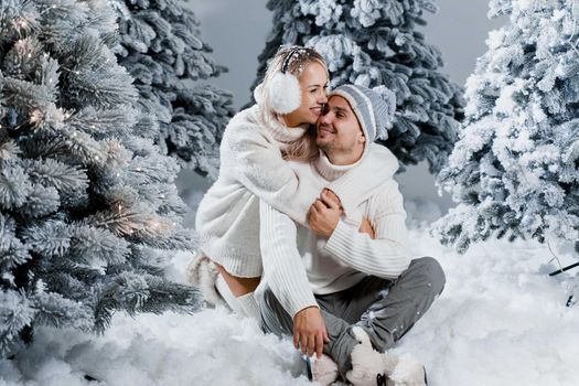 New year love story. Couple kisses and hugs. People weared wearing fur headphones, hats, white sweaters. Happy young couple hugs and kiss near christmas trees in winter day.