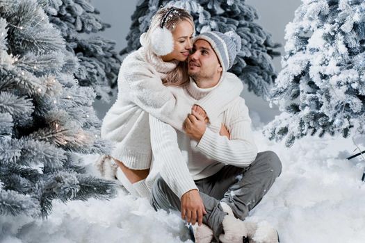 New year love story. Couple kisses and hugs. People weared wearing fur headphones, hats, white sweaters. Happy young couple hugs and kiss near christmas trees in winter day.