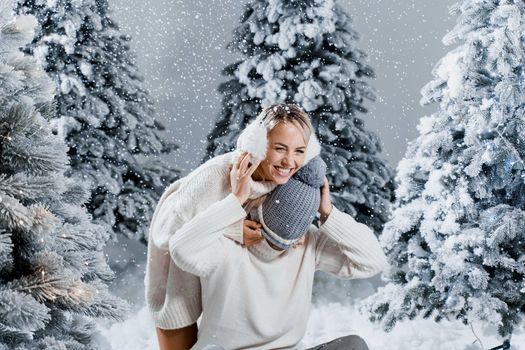 Winter love story with couple weared fur headphones, hats, white sweaters. Happy young couple hugs and kiss near christmas trees at the eve of new year celebration in winter day.