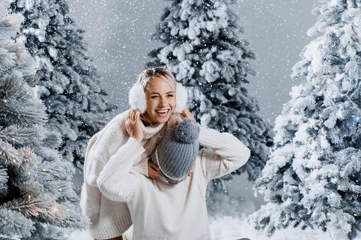 Winter love story with couple weared fur headphones, hats, white sweaters. Happy young couple hugs and kiss near christmas trees at the eve of new year celebration in winter day.