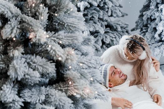 New year love story. Couple kisses and hugs. People weared wearing fur headphones, hats, white sweaters. Happy young couple hugs and kiss near christmas trees in winter day.