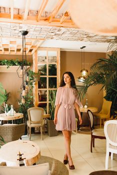 Young woman in soft pink dress is moving and looking into the camera. Beauty girl in cafe with big green palm tree