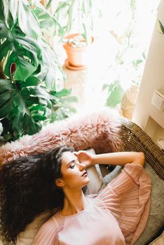 Dreamily woman is laying on the soft sofa and dream about summer vacation after quarantine. Beauty girl in cafe with green plants and big window. Stay at home