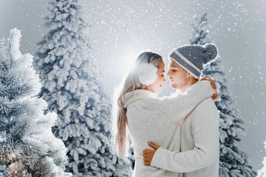 Falling snow and kisses. Happy young couple close-up hugs and kiss near christmas trees at the eve of new year celebration in winter day. Smiley man and woman weared white pullovers love each other