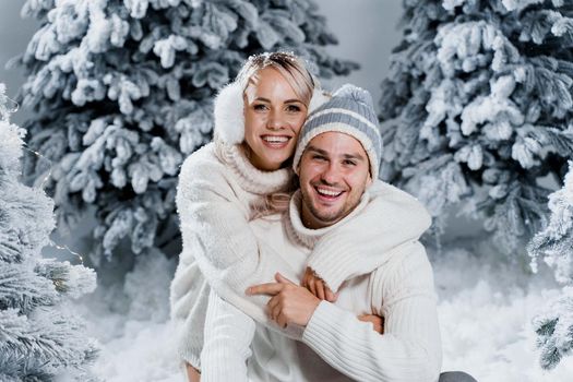 New year love story. Couple kisses and hugs. People weared wearing fur headphones, hats, white sweaters. Happy young couple hugs and kiss near christmas trees in winter day.