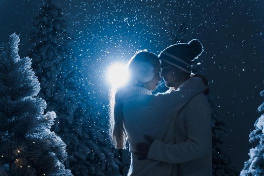 Falling snow and kisses with falling snow and dark blue background. Happy young couple weared white pullovers. hugs and kiss near christmas trees at the eve of new year celebration in winter day