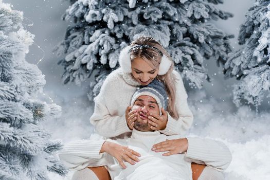 Falling snow and kisses. Happy young couple close-up hugs and kiss near christmas trees at the eve of new year celebration in winter day. Smiley man and woman weared white pullovers love each other