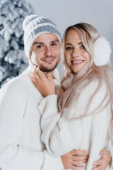 Winter love story with couple weared fur headphones, hats, white sweaters. Happy young couple hugs and kiss near christmas trees at the eve of new year celebration in winter day.