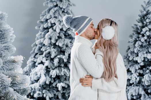 Couple kissing near christmass trees. Winter holidays. Love story of young couple weared white pullovers. Happy man and young woman hug each other.