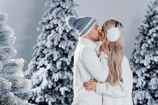 Winter love story with couple weared fur headphones, hats, white sweaters. Happy young couple hugs and kiss near christmas trees at the eve of new year celebration in winter day.