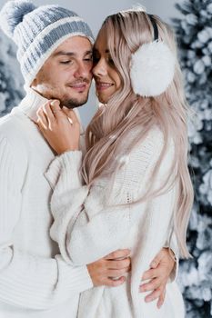 Winter love story with couple weared fur headphones, hats, white sweaters. Happy young couple hugs and kiss near christmas trees at the eve of new year celebration in winter day.