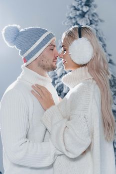 Winter love story with couple weared fur headphones, hats, white sweaters. Happy young couple hugs and kiss near christmas trees at the eve of new year celebration in winter day.