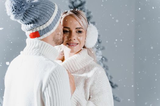 Happy young couple hugs and smiles and snow falls near christmas trees at the eve of new year celebration in winter day. Smiley man and woman weared white pullovers love each other