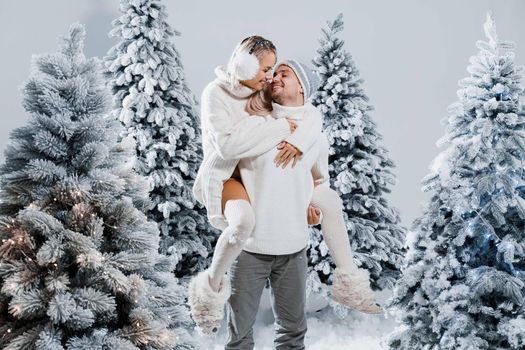 Couple kiss and hug. Man holds girl near christmas trees in winter day. New year celebration. People weared wearing fur headphones, hats, white sweaters.