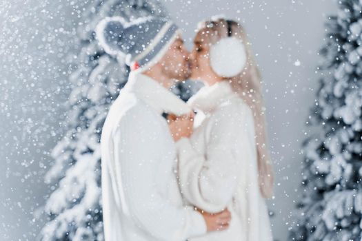 Happy young couple hugs and smiles and snow falls near christmas trees at the eve of new year celebration in winter day. Smiley man and woman weared white pullovers love each other