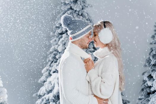 Happy young couple hugs and smiles and snow falls near christmas trees at the eve of new year celebration in winter day. Smiley man and woman weared white pullovers love each other