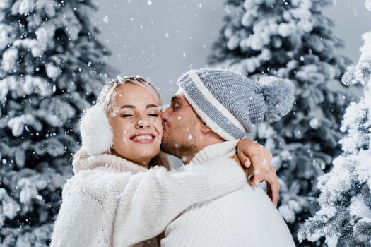 Falling snow and kisses. Happy young couple close-up hugs and kiss near christmas trees at the eve of new year celebration in winter day. Smiley man and woman weared white pullovers love each other