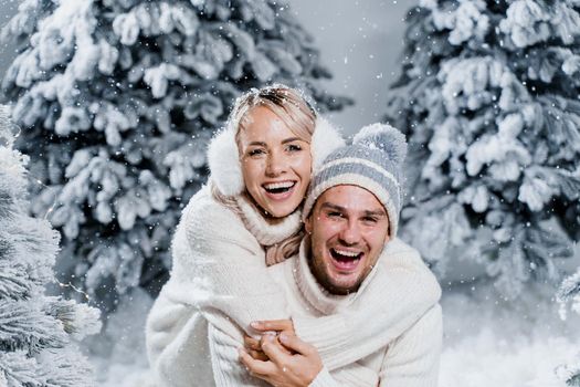 New year love story. Couple kisses and hugs. People weared wearing fur headphones, hats, white sweaters. Happy young couple hugs and kiss near christmas trees in winter day.