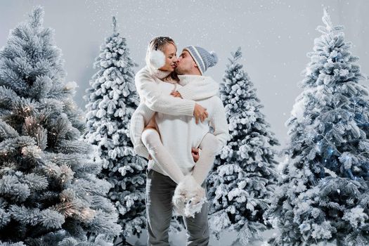 Couple kiss and hug. Man holds girl near christmas trees in winter day. New year celebration. People weared wearing fur headphones, hats, white sweaters.