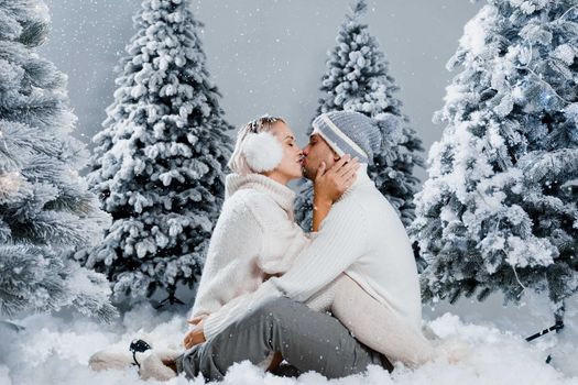 Falling snow and kisses. Happy young couple close-up hugs and kiss near christmas trees at the eve of new year celebration in winter day. Smiley man and woman weared white pullovers love each other