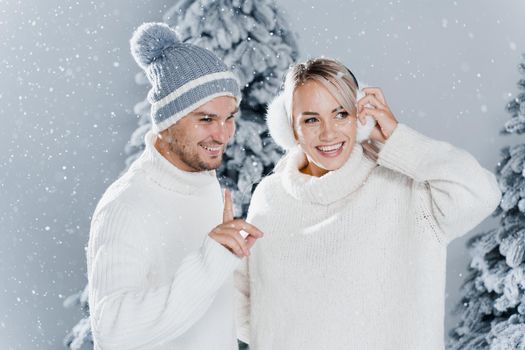 Couple couple laughing and having fun while snow falls near christmass trees. Winter holidays. Love story of young couple weared white pullovers. Happy man and young woman hug each other.